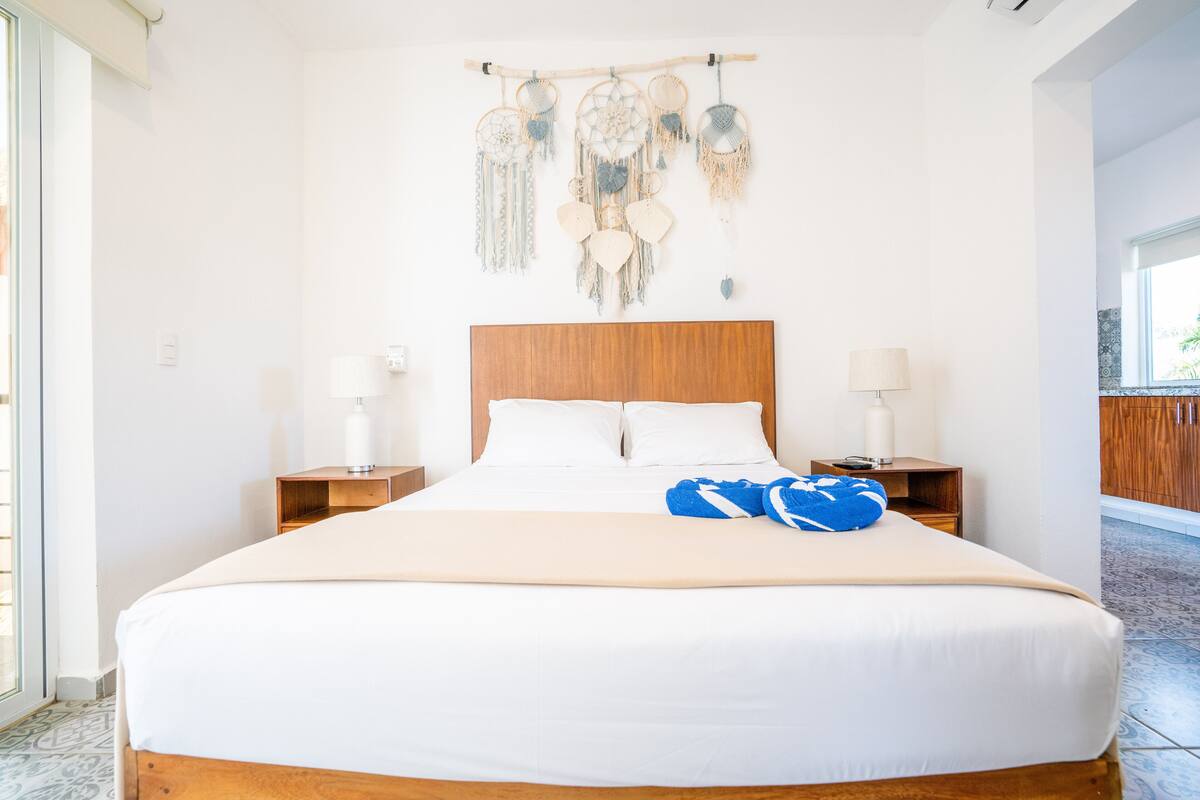 Ein ordentlich bezogenes Bett mit weißen Leinen, einem Holzkopfende und einem blauen Kopfkissen. Es gibt zwei Nachttische mit Lampen und eine an der Wand befestigte Makramee-Dekoration über dem Bett.