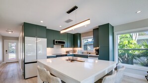 A modern kitchen with a central island, white bar stools, and a view of greenery outside.