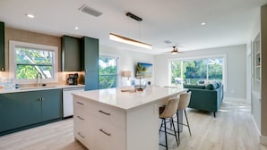 A modern kitchen with a central island, a dining area with a green sofa, and large windows offering a view of greenery.