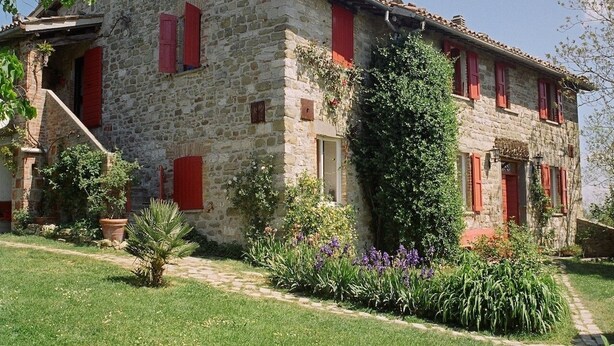 A stone building with red shutters, a garden with various plants, and a pathway leading to the entrance.