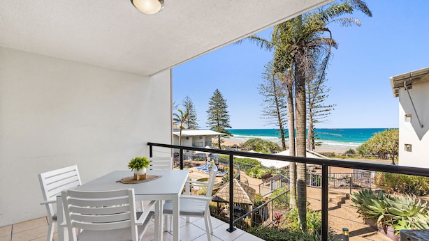 A balcony with a view of the beach and ocean, white chairs, a small table, and a potted plant.