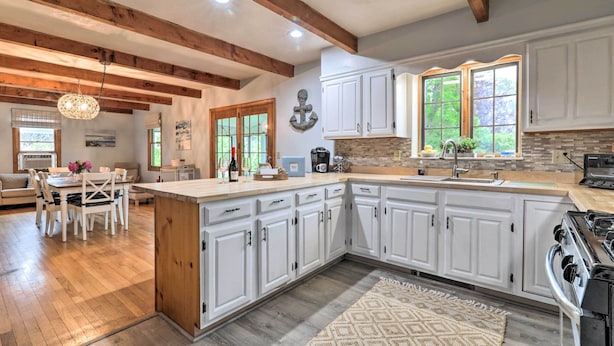 A spacious kitchen with white cabinets, a large island, and a stone backsplash. There is a dining area with a table and chairs, and a view of the outdoors through large windows. - Walk to Main Street: Cottage w/ Sunroom in Hyannis (Barnstable)