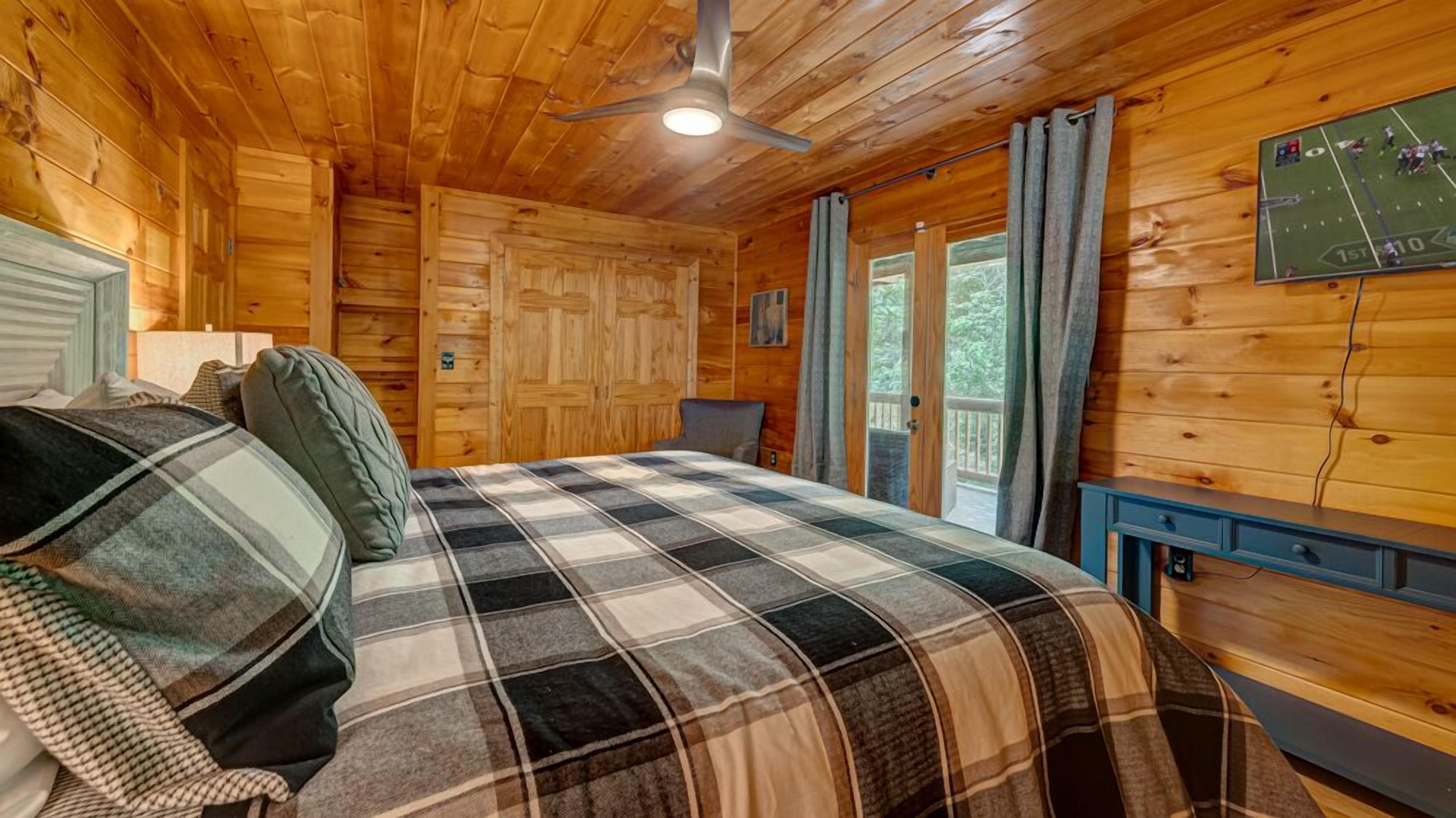 Une chambre dans un chalet en bois, avec un lit, un ventilateur de plafond, un téléviseur fixé au mur et une fenêtre avec des rideaux.
