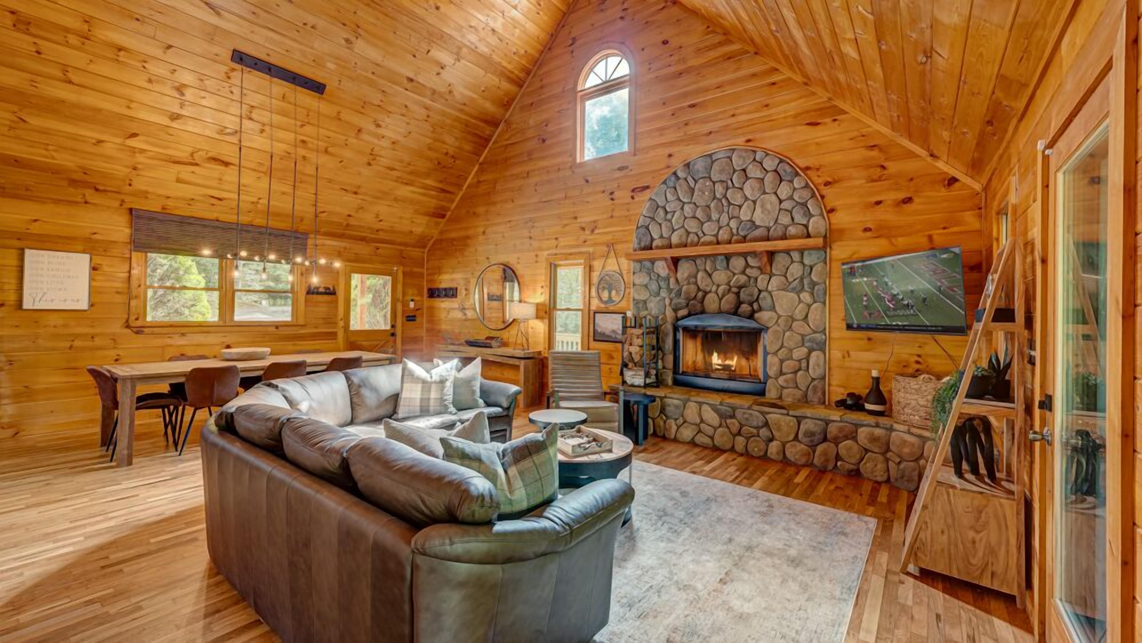 L’intérieur d’un chalet confortable, avec une cheminée en pierre, un canapé en cuir, un coin repas et un téléviseur.
