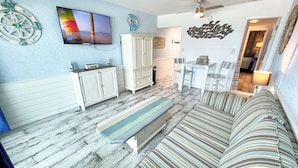Una sala de estar con un sofá a rayas, un televisor, un mueble blanco y una zona de comedor con una mesa y sillas.