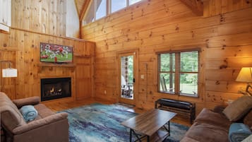 A cozy log cabin interior with a fireplace, a television mounted on the wall, and a large window providing natural light.