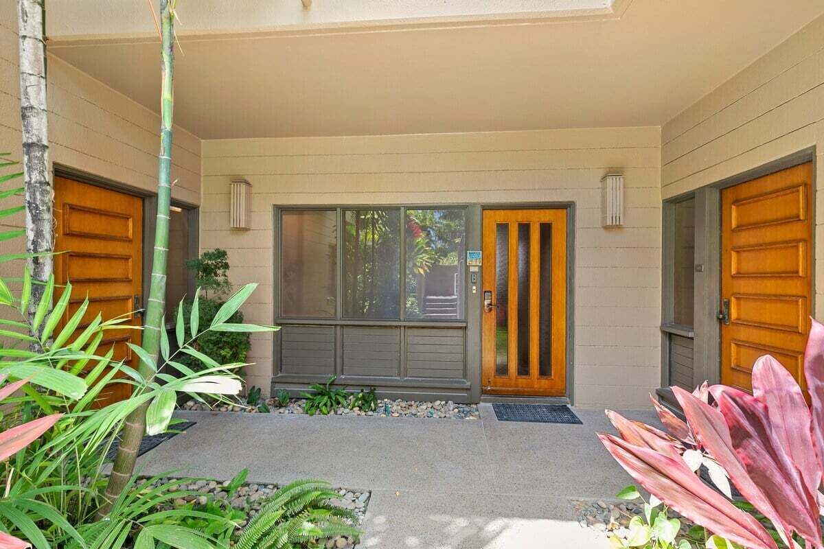 Une entrée d’immeuble avec deux portes en bois, un petit jardin agrémenté de plantes et une fenêtre donnant sur un espace verdoyant.