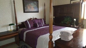A bedroom with a four-poster bed, a nightstand, a desk, a chair, and a large window.