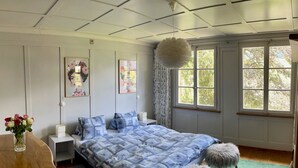 Een slaapkamer met een bed, nachtkastjes, een dressoir, een hanglamp en een raam met uitzicht op bomen.