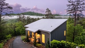 Une maison moderne avec un toit en métal, entourée d’une forêt dense et de montagnes brumeuses.
