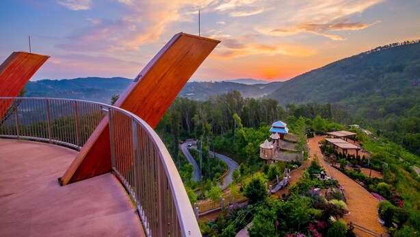 Une vue magnifique depuis un balcon donnant sur un jardin luxuriant et un bâtiment traditionnel à toit bleu, le tout se détachant sur un arrière-plan de collines ondoyantes et d’un ciel au coucher du soleil.