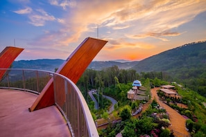A scenic view from a balcony overlooking a lush garden and a traditional building with a blue roof, set against a backdrop of rolling hills and a sunset sky. - Private modern indoor pool cabin | Dollywood (Sevierville)
