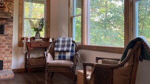 Habitación acogedora con pisos de madera, chimenea de ladrillo y ventanas amplias con vista a árboles. Hay dos sillones de mimbre y una mesita con un florero.