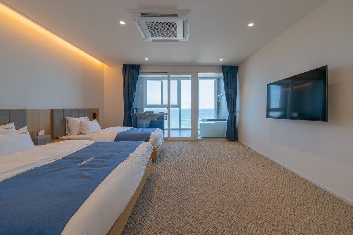 A hotel room with two beds, a large TV, and a balcony with a view of the sea.