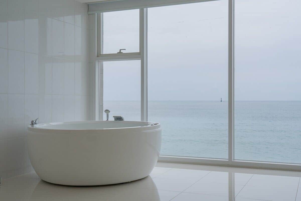 A modern bathroom with a large bathtub, white tiles, and a view of the sea through large windows.
