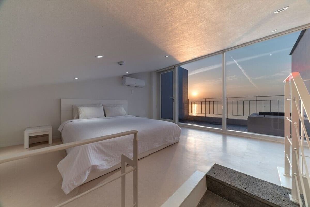 A modern bedroom with a large bed, a small bedside table, a nightstand, and a view of the sunset through large windows.