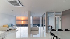 A modern living room with a sofa, a dining table, and a view of the sunset through large windows. - Daytona Pool Villa (Pohang)