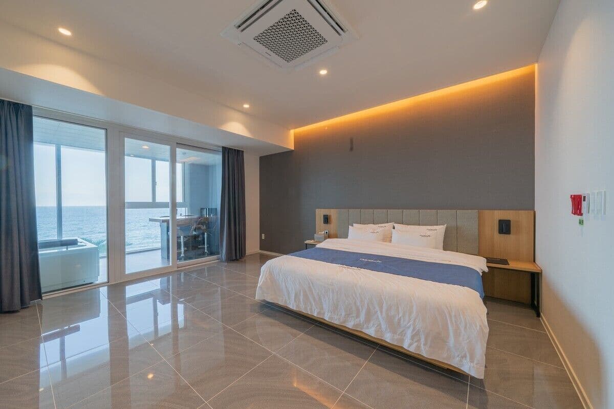A modern hotel room with a large bed, a sliding glass door leading to a balcony with a view of the sea, and a well-lit room with a ceiling vent.