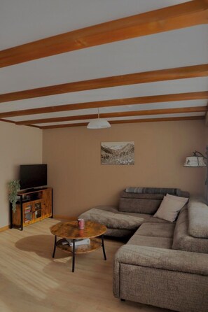Una sala de estar con un sofá seccional, una mesa de centro redonda, un televisor sobre un mueble de madera y un cuadro en la pared.