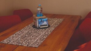 Un coin repas avec une table en bois, des chaises à coussins rouges, une bouteille d’eau et un tableau représentant une vache accroché au mur.