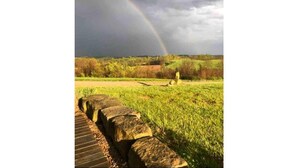 Ein steiniger Weg führt zu einer grünen Fläche mit einer Statue, unter einem bewölkten Himmel mit einem Regenbogen.