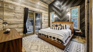 A bedroom with a wooden ceiling, a bed with striped bedding, a nightstand, and a window with a view of trees. - BRAND NEW! Smokey Pines |  BOHO Modern + Chic Contemporary | Sleeps 12 (Broken Bow)