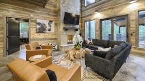 A log cabin interior with a stone fireplace, a mounted TV, and a cozy seating area with leather sofas and a coffee table. - BRAND NEW! Smokey Pines |  BOHO Modern + Chic Contemporary | Sleeps 12 (Broken Bow)