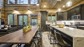 A spacious kitchen with dark cabinets, a large island with bar stools, and a dining area with a long table and chairs. - BRAND NEW! Smokey Pines |  BOHO Modern + Chic Contemporary | Sleeps 12 (Broken Bow)