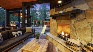 A cozy outdoor seating area with a stone fireplace, a wooden deck, and a swing set visible through the trees. - BRAND NEW! Smokey Pines |  BOHO Modern + Chic Contemporary | Sleeps 12 (Broken Bow)