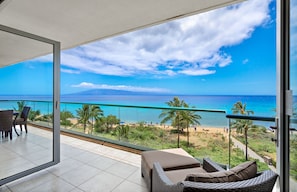 A balcony with a view of the beach and ocean, wicker furniture, and a glass railing.