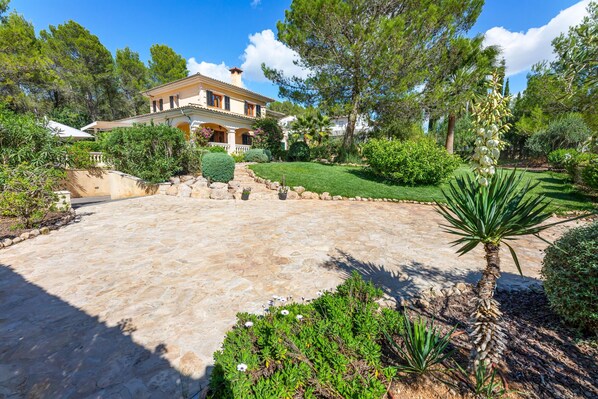 A Mediterranean-style villa with a stone courtyard, lush greenery, and a prominent palm tree.