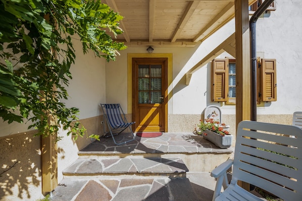 A stone-paved entrance with a wooden door, a blue chair, a white chair, and a potted plant with flowers.