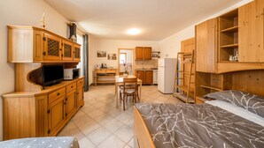 A compact room with a bed, a desk, a television, wooden cabinets, and a kitchenette.