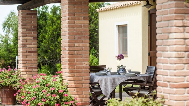 A covered patio with a brick column, a table set with plates, glasses, and a pitcher, surrounded by potted plants and a building in the background. - Holiday Home "Asfodeli" with Wi-Fi, A/C & Garden (Teulada)