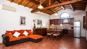 Un soggiorno con divano, tavolo da pranzo, sedie, angolo cottura con pensili ed elettrodomestici, e un alto soffitto in legno.