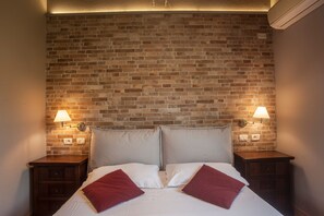 A hotel room with a brick-patterned headboard, two bedside tables with lamps, a bed with white linens and red pillows, and a nightstand with a drawer.