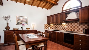A kitchen with wooden cabinets, a dining table with chairs, a hanging light fixture, and a window with a view of greenery. - Holiday Home "Asfodeli" with Wi-Fi, A/C & Garden (Teulada)