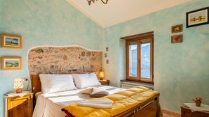 Une chambre à coucher avec une tête de lit en pierre, un lit avec une literie blanche et jaune, une table de chevet avec une lampe, un cadre de fenêtre en bois et une vue sur l’extérieur.