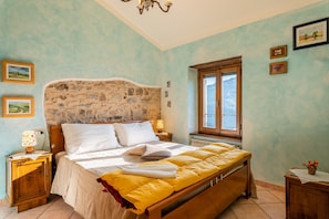 Quarto com cabeceira de pedra, cama com roupa de cama branca e amarela, mesa de cabeceira com abajur, moldura de janela de madeira e vista para o exterior.