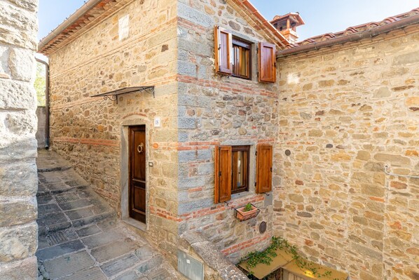 Un bâtiment en pierre avec une porte en bois, une fenêtre avec des volets et un petit jardinière posée sur le sol.