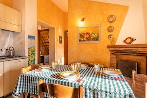 Un coin repas avec une nappe à carreaux, une cheminée et un tableau représentant des fruits accroché au mur.