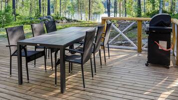 Una terraza de madera con una mesa y sillas, una parrilla y vistas a un bosque y un lago.