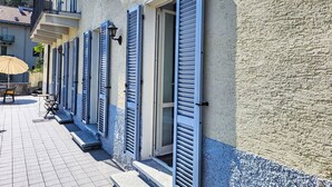 A building with blue shutters, a tiled patio, and a small outdoor seating area. - Casa Stella App. 1 by Interhome (Brissago)