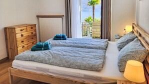 A double bed with patterned bedding, a wooden dresser, a nightstand with a lamp, and a view of a balcony with plants and a mountain. - Casa Stella App. 1 by Interhome (Brissago)