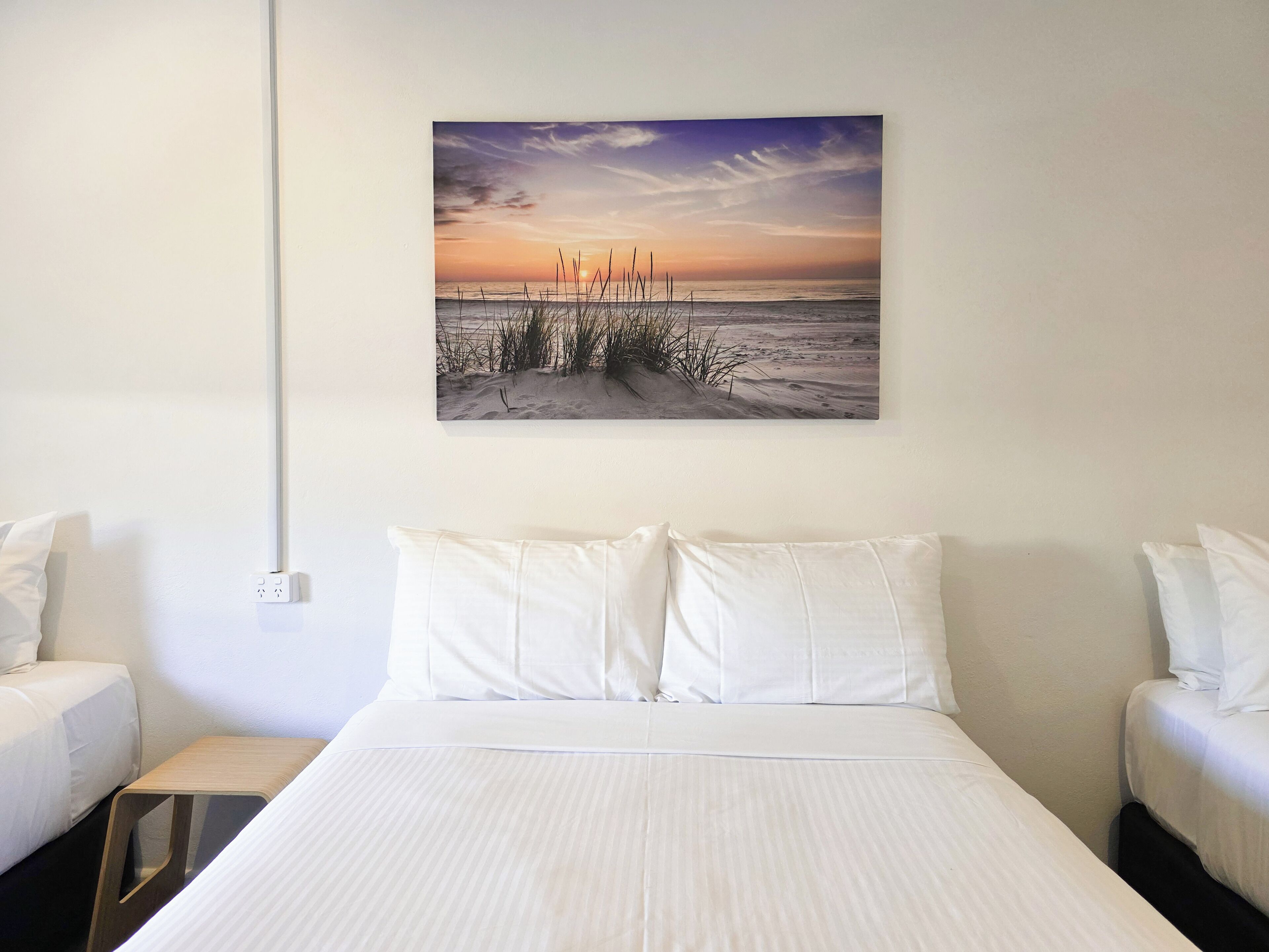 A hotel room with a large bed, two white pillows, a wooden nightstand, and a wall-mounted painting of a beach sunset.