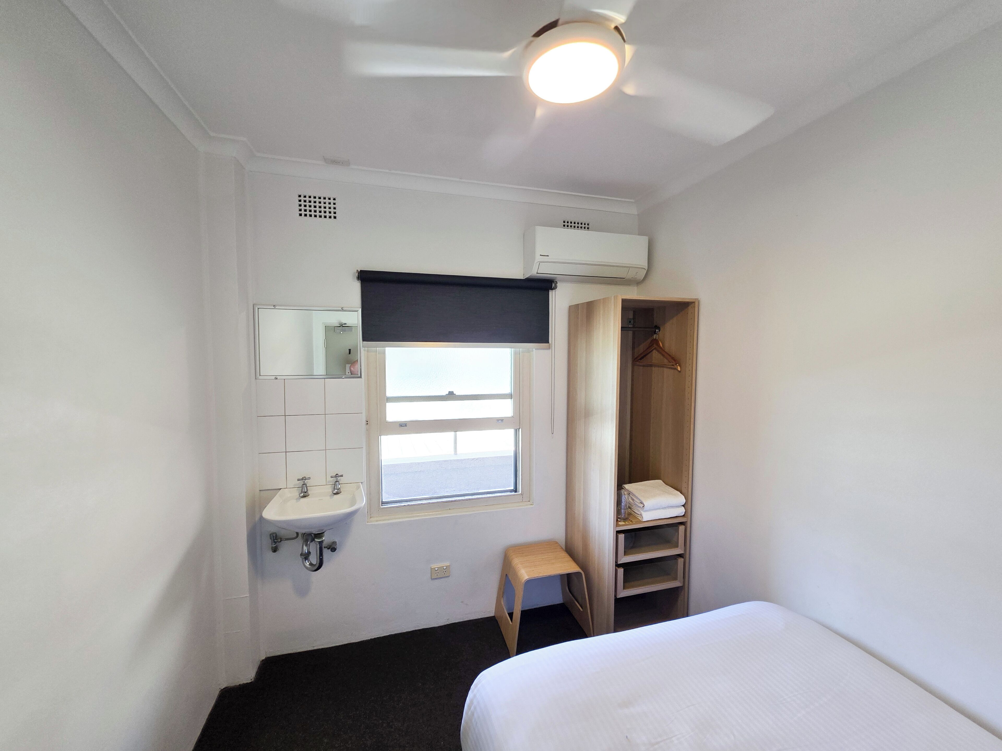 A small, clean room with a bed, a window, a mirror, a sink, and a wardrobe.