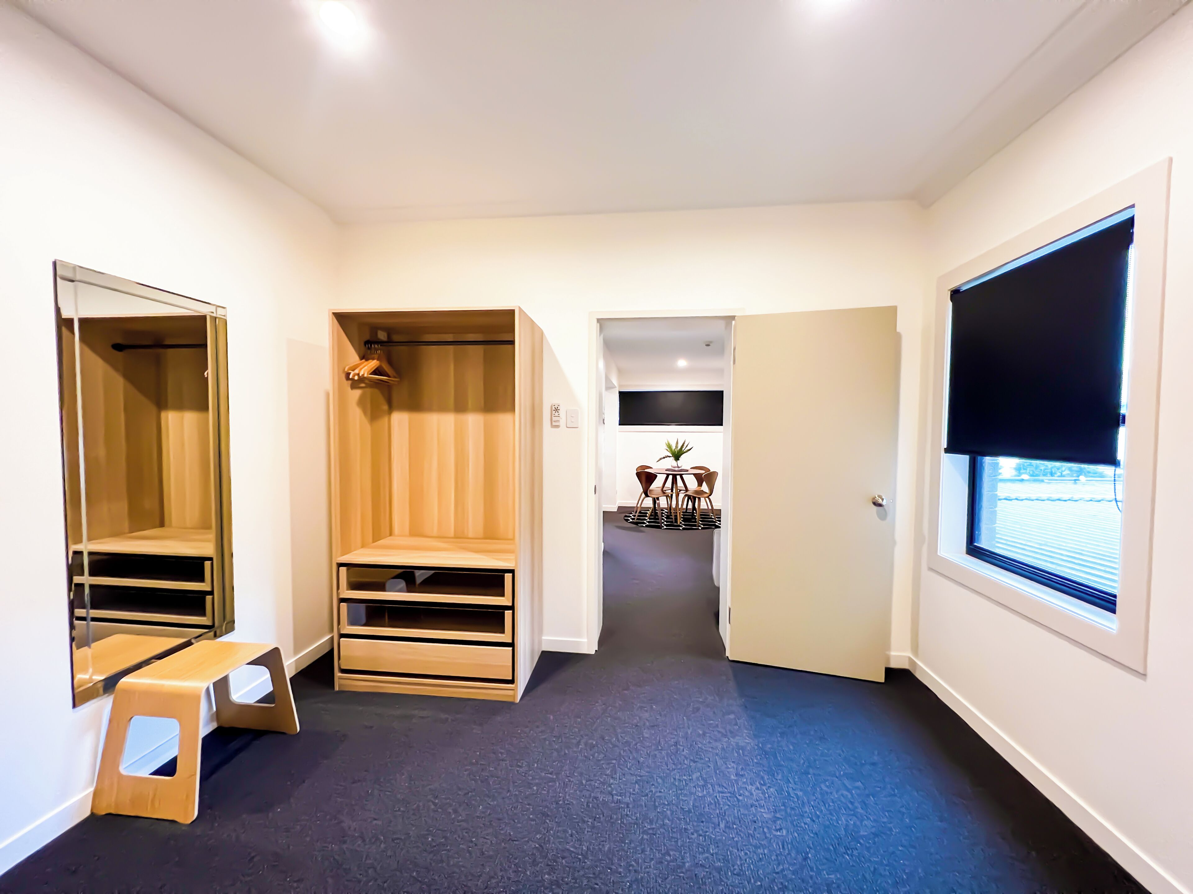A hallway with a wooden closet, a small stool, a dining area with a table and chairs, and a window with blinds.