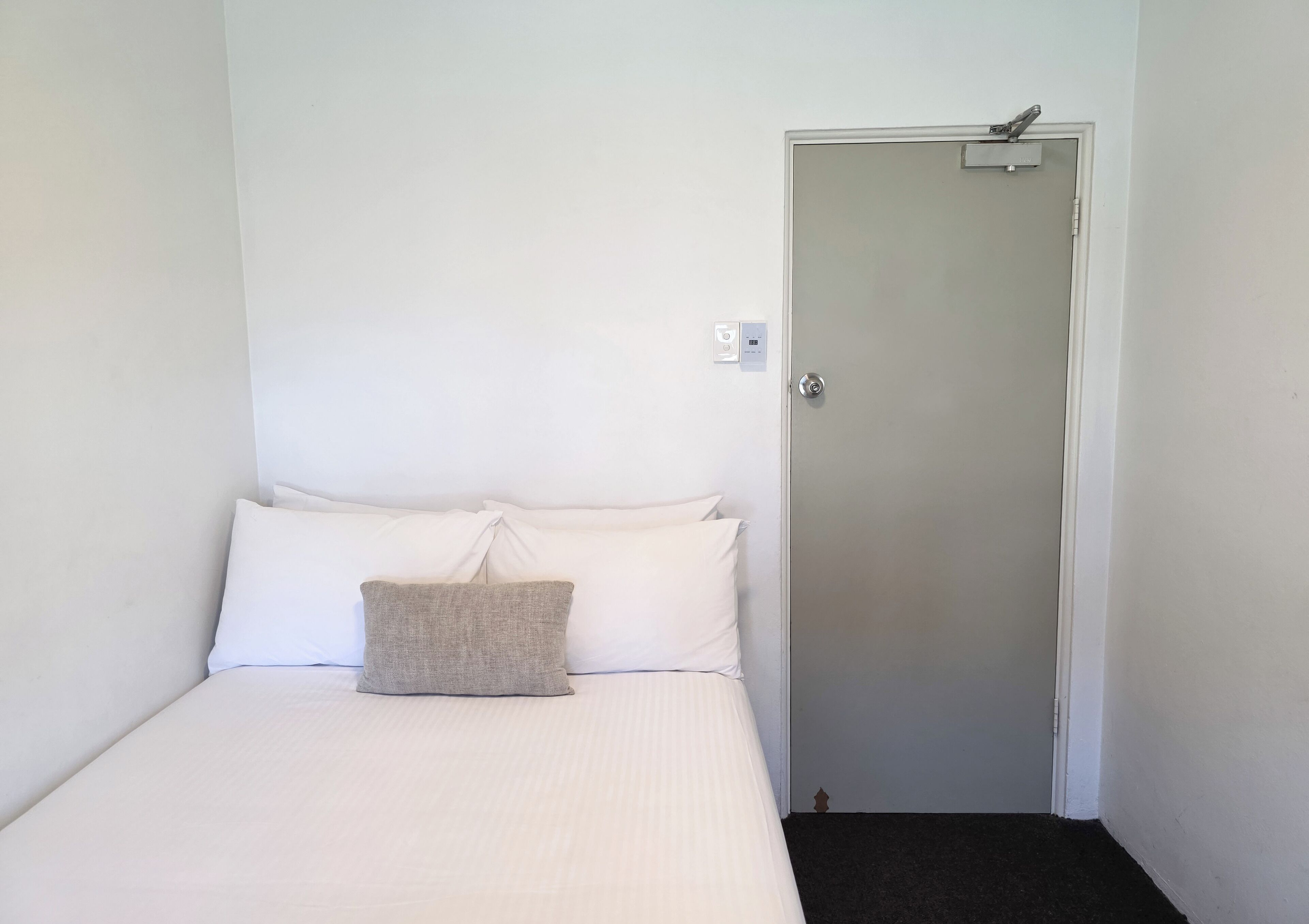 A single bed with white bedding and a grey pillow, a door with a handle, and a light switch on the wall.