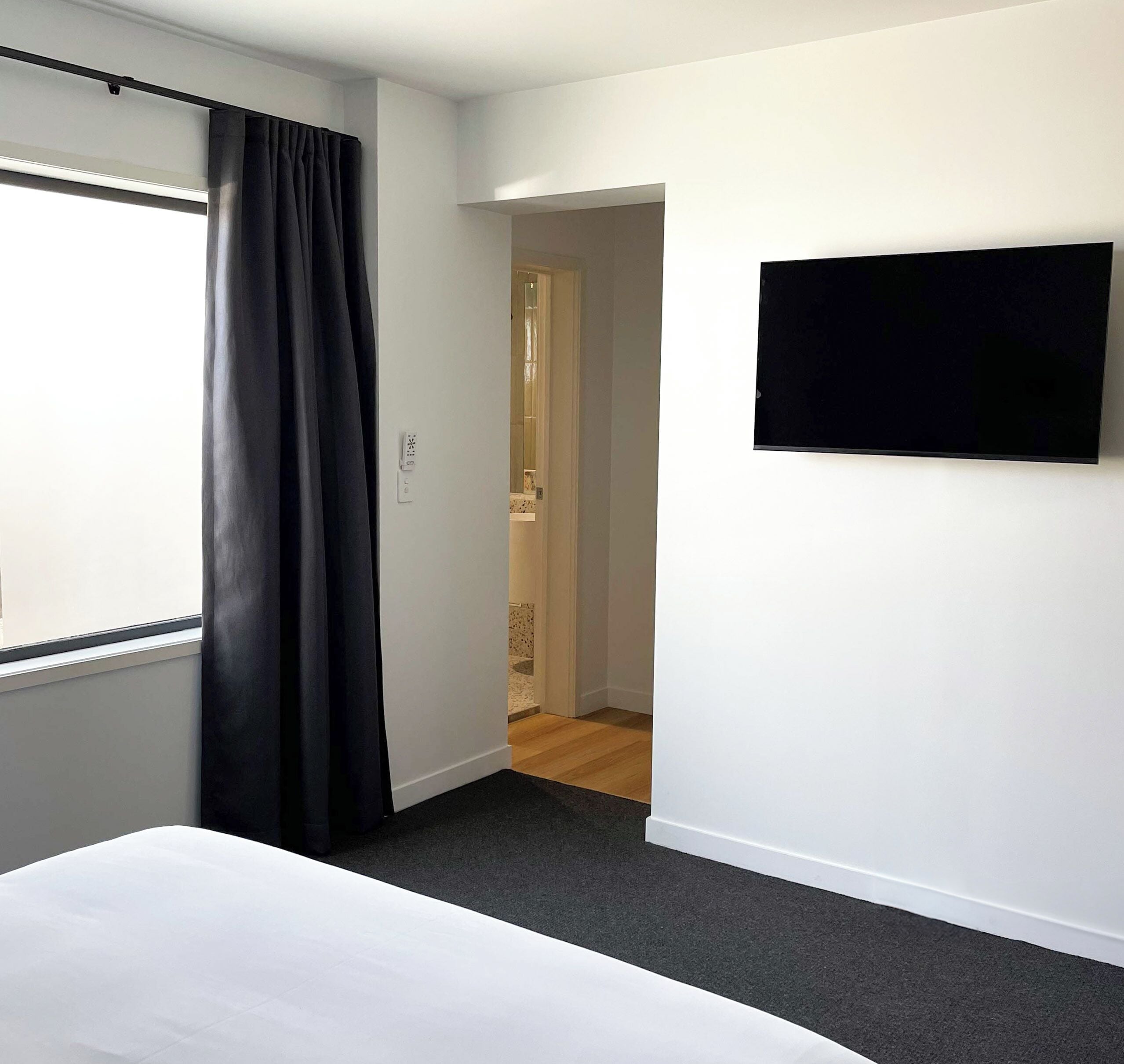 A hotel room with a bed, a TV mounted on the wall, a window with curtains, and a door leading to another room.