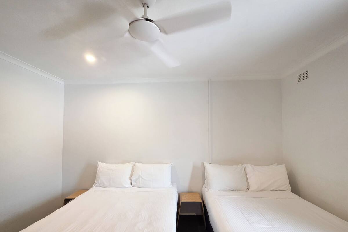 Una habitación sencilla y limpia con dos camas individuales, un ventilador de techo y una mesita de noche de madera.
